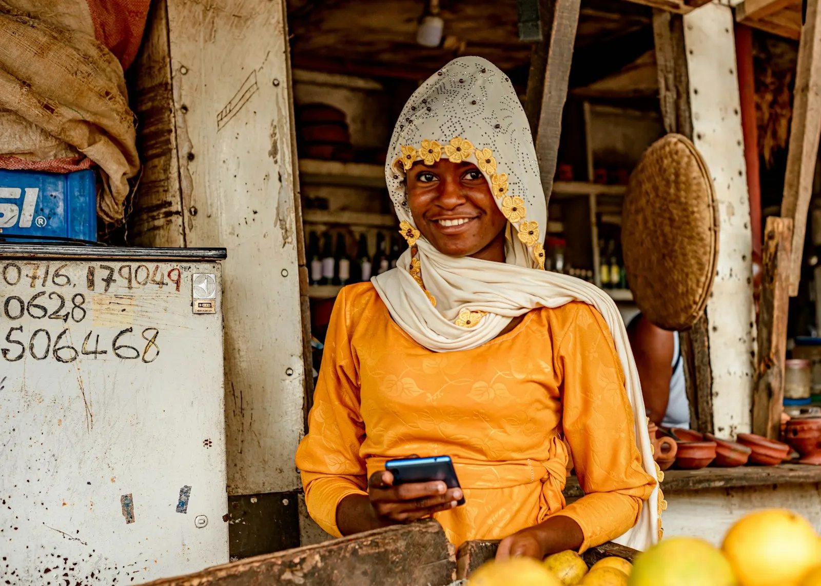 A smiling shopkeeper holding a phone at her kiosk, phone numbers written on the wall beside her