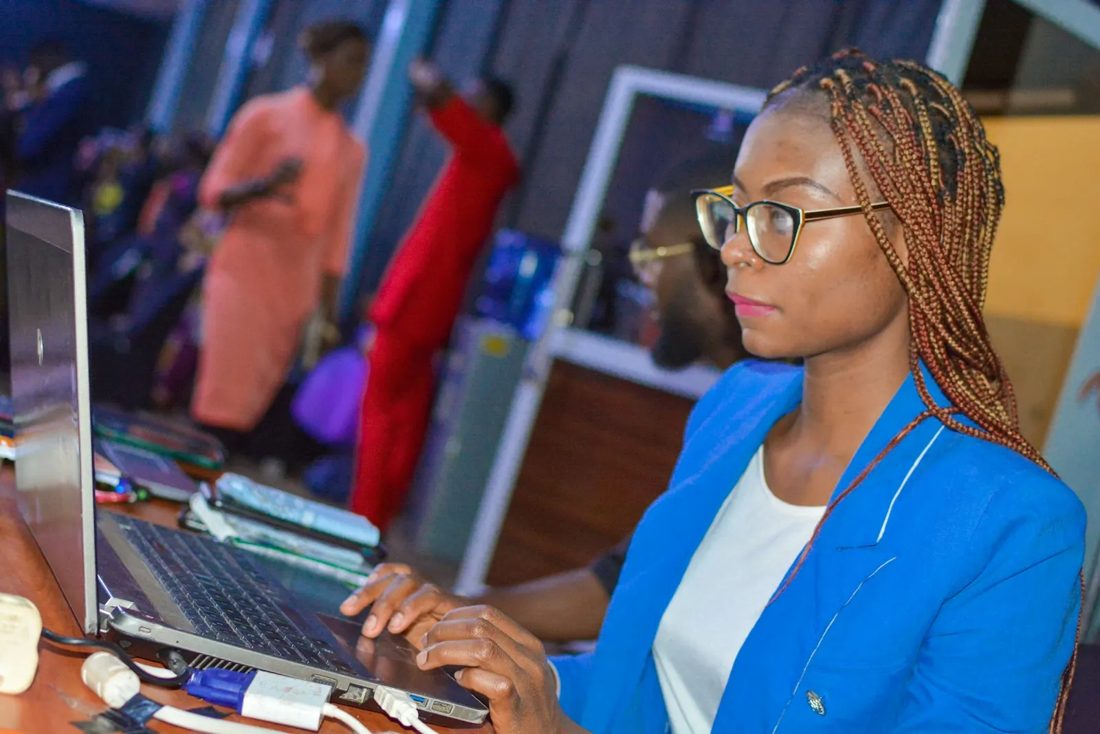 A woman in glasses working on a laptop in a busy office